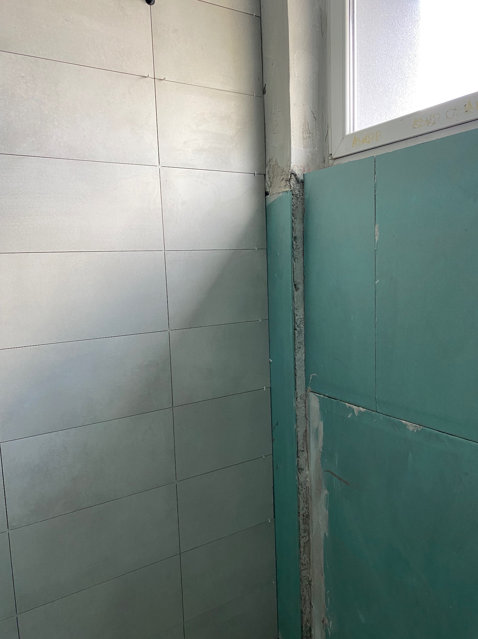 Ściana z szarymi płytkami i zielonymi płytami gipsowo-kartonowymi, widoczne łączenia i narożnik przy oknie. Prace wykończeniowe w trakcie realizacji.