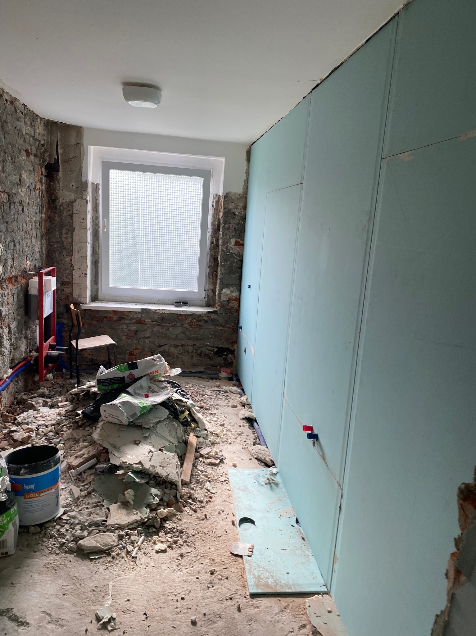 Montaż płyt karton-gips na ścianie w trakcie remontu. Widoczne worki z zaprawą, gruz i instalacja sanitarna. Okno z kratką w tle.
