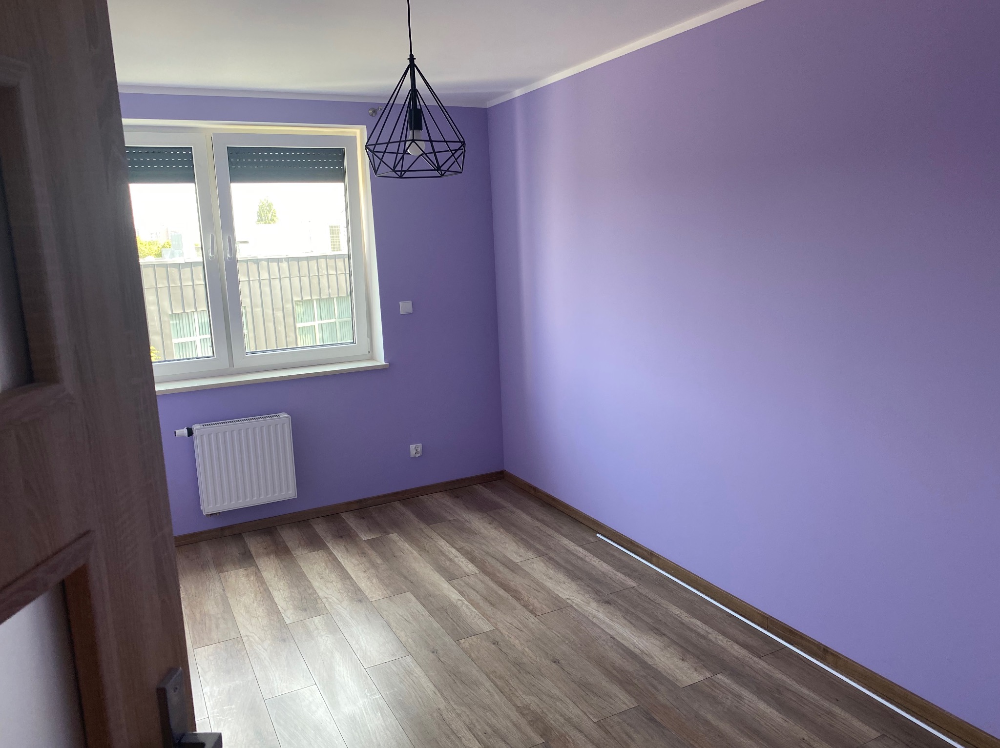 Pomalowane na fioletowo puste wnętrze pokoju z drewnianą podłogą, białym grzejnikiem i geometryczną lampą wiszącą. Widok zza uchylonych drzwi.