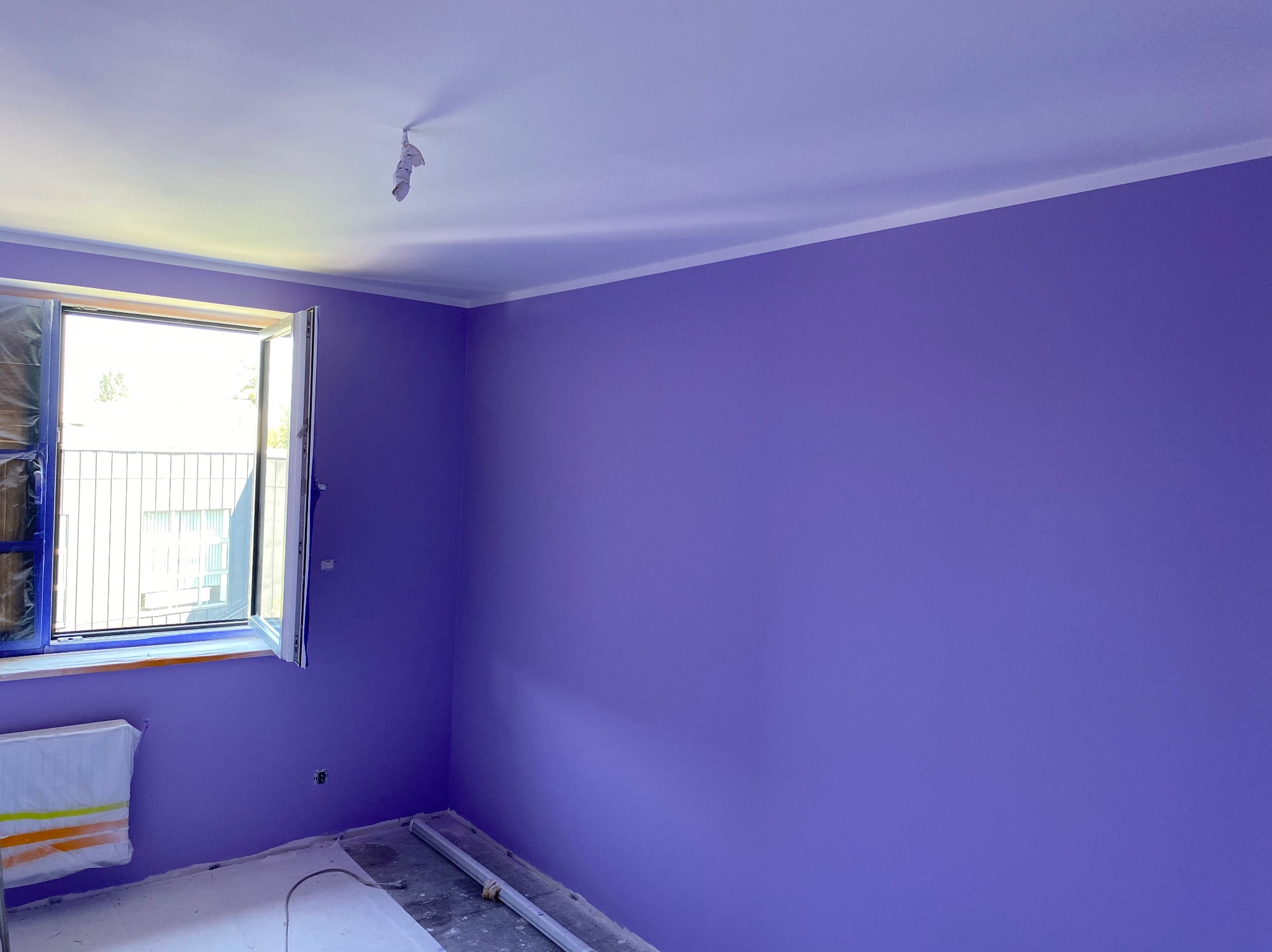 Pomieszczenie po malowaniu ścian na fioletowo. Okno zabezpieczone folią, grzejnik zakryty. Widoczne listwy przypodłogowe i fragment sufitu z kablem.