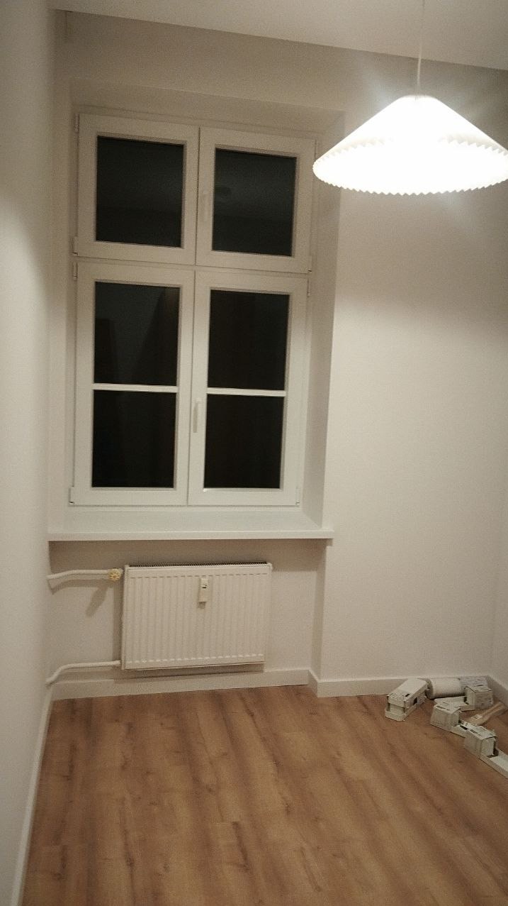 Jasne wnętrze po remoncie: białe ściany, okno z grzejnikiem, drewniana podłoga. Widoczne listwy przypodłogowe i lampa sufitowa. Minimalistyczny design.