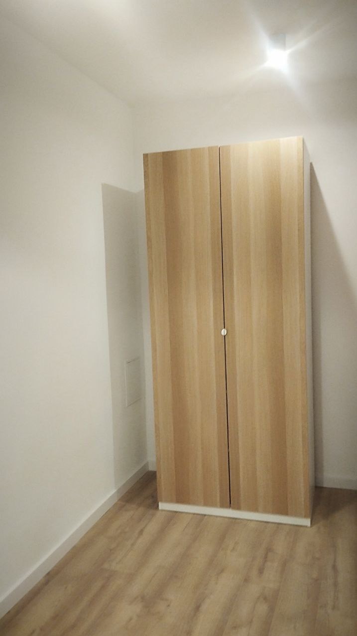 Jasne, drewniane drzwi szafy wnękowej w minimalistycznym wnętrzu z panelami podłogowymi. Ściany pomalowane na biało, widoczne gniazdko elektryczne.