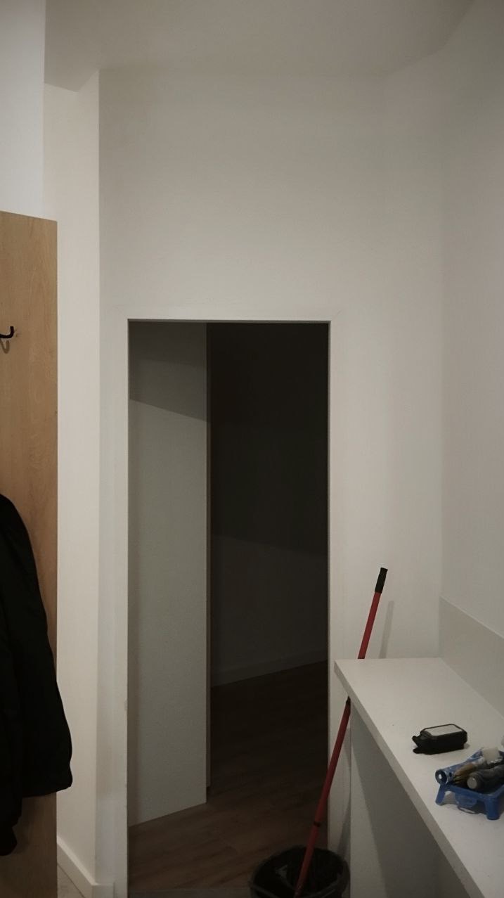 Świeżo pomalowane na biało ściany w korytarzu z widocznym wejściem do innego pomieszczenia. Na pierwszym planie akcesoria malarskie i wiadro z mopem.