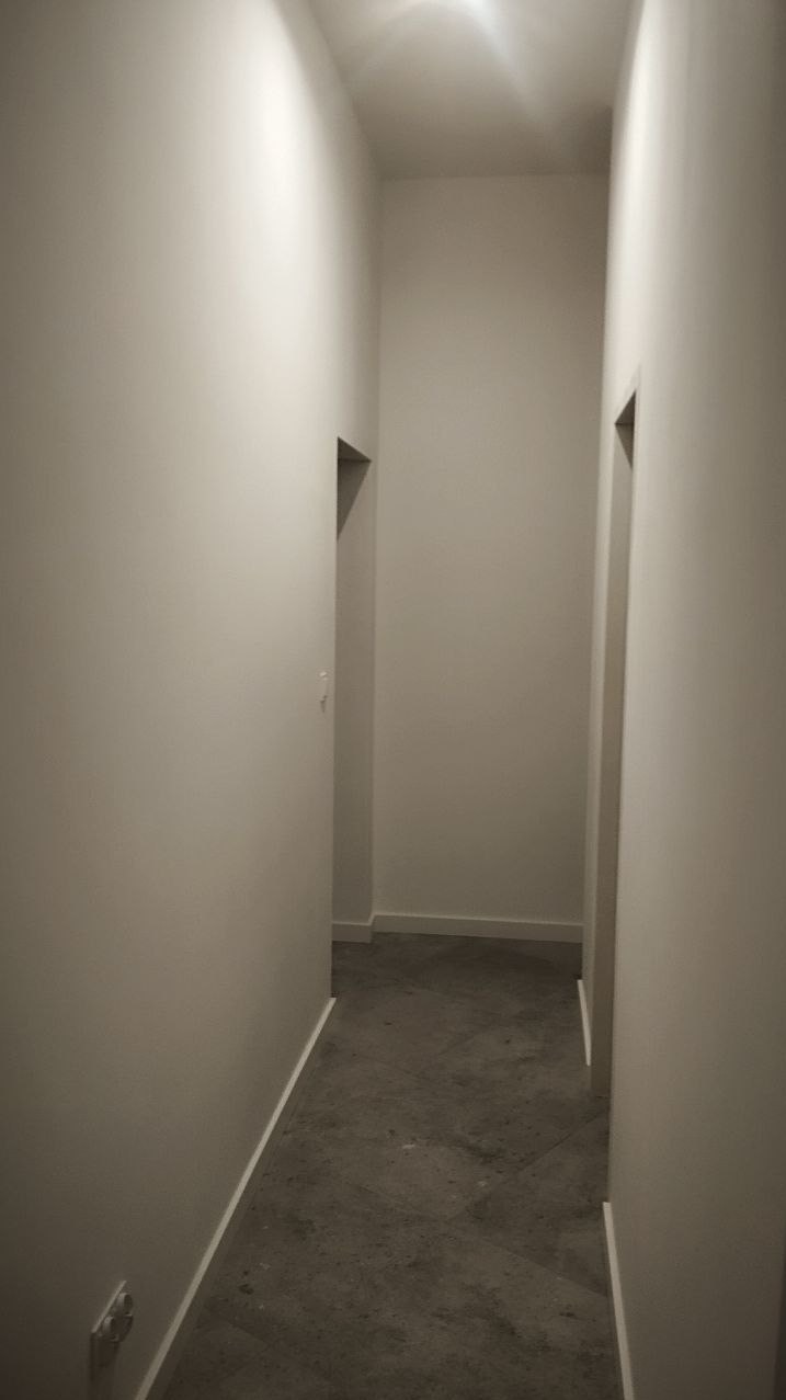 Wąski korytarz po remoncie z szarymi płytkami na podłodze i świeżo pomalowanymi na biało ścianami. Widoczne drzwi po obu stronach korytarza i listwy przypodłogowe.