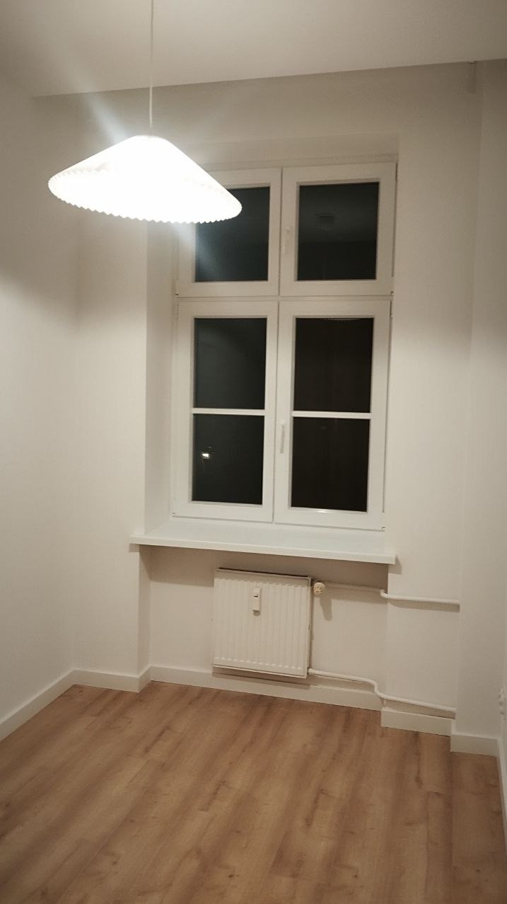 Białe, odświeżone wnętrze pokoju po remoncie: drewniana podłoga, okno z białymi ramami, białe ściany i sufit, oraz lampa wisząca. Widoczny grzejnik pod oknem.