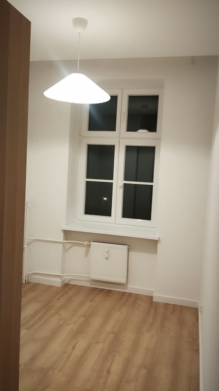 Jasne wnętrze po remoncie: białe ściany, okno z białymi ramami, drewniana podłoga i nowoczesna lampa wisząca. Minimalistyczny design, świeżo po malowaniu.