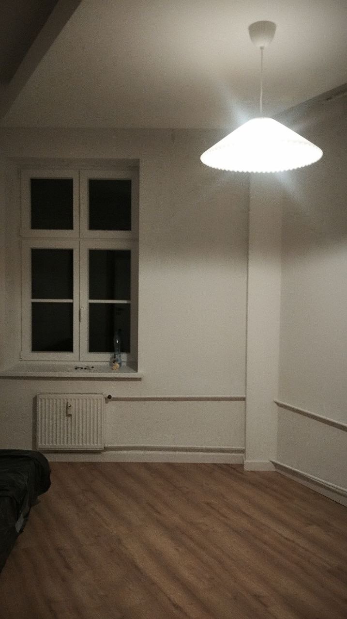 Pomalowane na biało wnętrze pokoju z drewnianą podłogą, oświetlone lampą sufitową. Widoczne okno z grzejnikiem i dekoracyjnymi listwami przypodłogowymi.