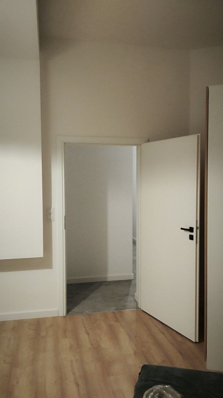 Jasne wnętrze po remoncie: świeżo pomalowane ściany, drewniana podłoga i otwarte drzwi do kolejnego pomieszczenia z szarą podłogą. Minimalistyczny design.