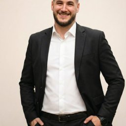 AG NIERUCHOMOŚCI NOVA SPÓŁKA Z OGRANICZONA ODPOWIEDZIALNOŚCIĄ - Uśmiechnięty mężczyzna w czarnym garniturze i białej koszuli, z rękami w kieszeniach, prezentuje się profesjonalnie na jasnym tle. Widoczny zegarek na nadgarstku.