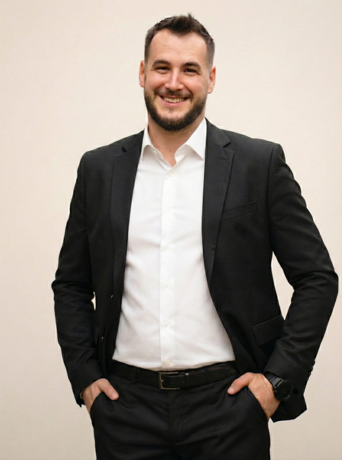 Uśmiechnięty mężczyzna w czarnym garniturze i białej koszuli, z rękami w kieszeniach, prezentuje się profesjonalnie na jasnym tle. Widoczny zegarek na nadgarstku.