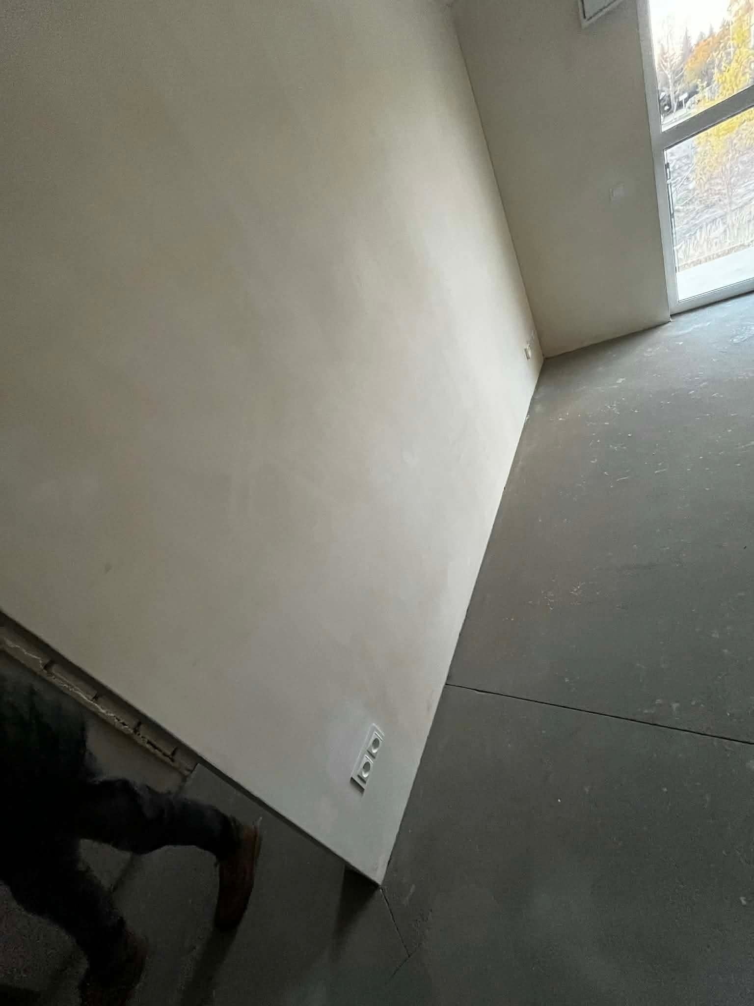 Gładka, jasna ściana po malowaniu w nowym mieszkaniu, widoczne gniazdka elektryczne i fragment okna z widokiem na zewnątrz. Szara podłoga w surowym stanie.