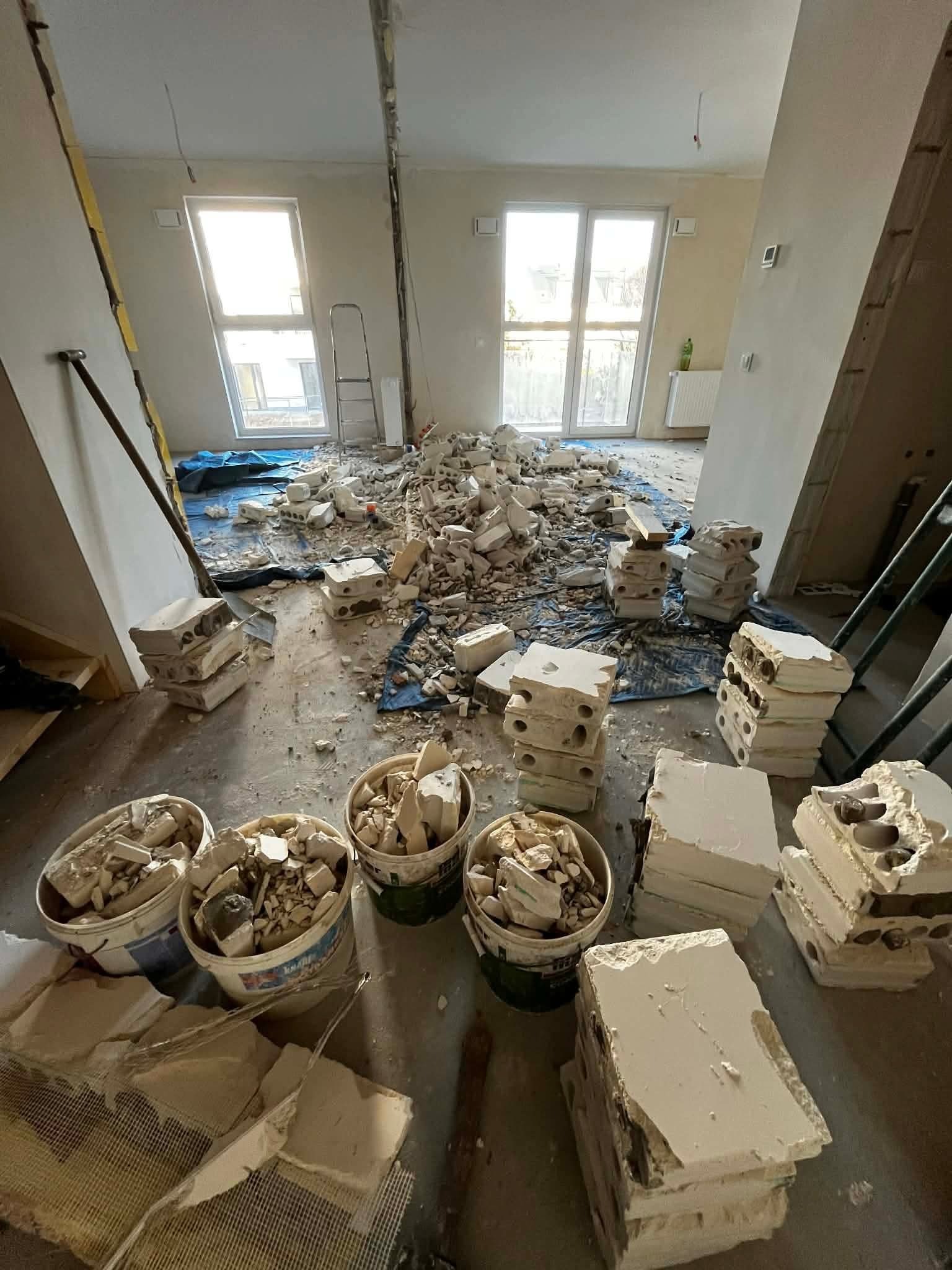Wnętrze po demontażu ściany: gruz w wiadrach i ułożone fragmenty muru na podłodze, zabezpieczonej folią. Widoczne okna i drabina w tle.