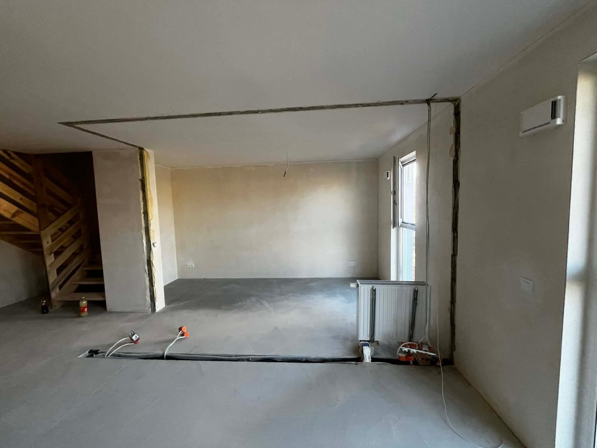 Wnętrze w trakcie remontu: widoczne bruzdy w ścianach pod instalacje elektryczne, wylewka podłogowa, grzejnik i schody drewniane. Prace przygotowawcze do malowania.