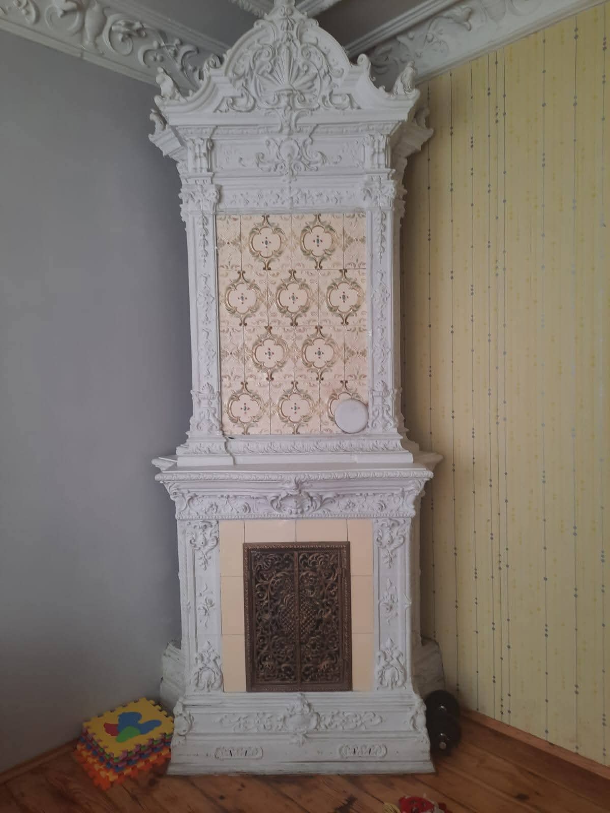 Ozdobny, biały piec kaflowy z dekoracyjnymi elementami sztukaterii i wzorzystymi kaflami, stojący w rogu pokoju z szarą i żółtą ścianą.
