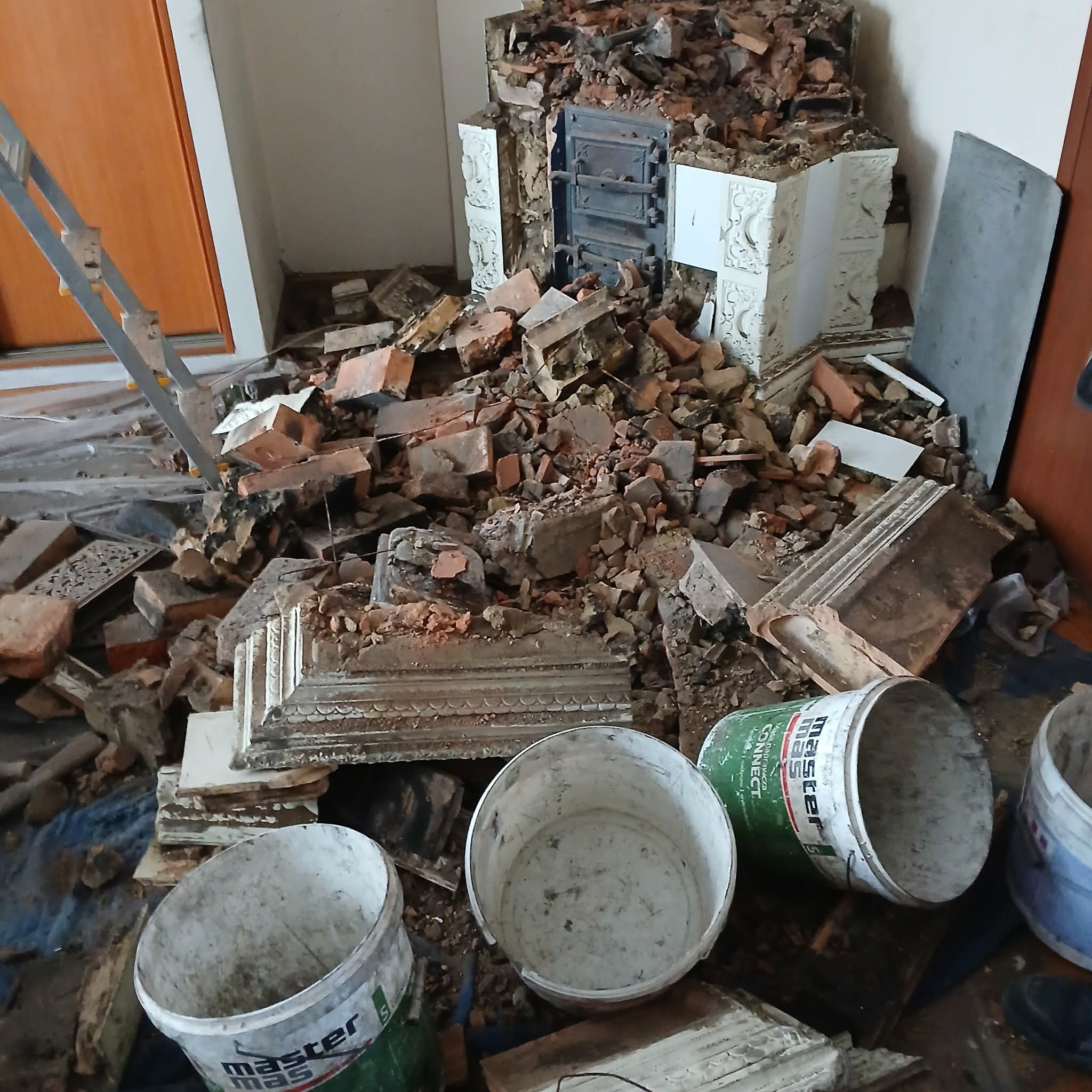 Demontaż pieca kaflowego w pomieszczeniu. Widoczne gruz, cegły, fragmenty kafli i puste wiadra po farbie. Drabina oparta o ścianę.