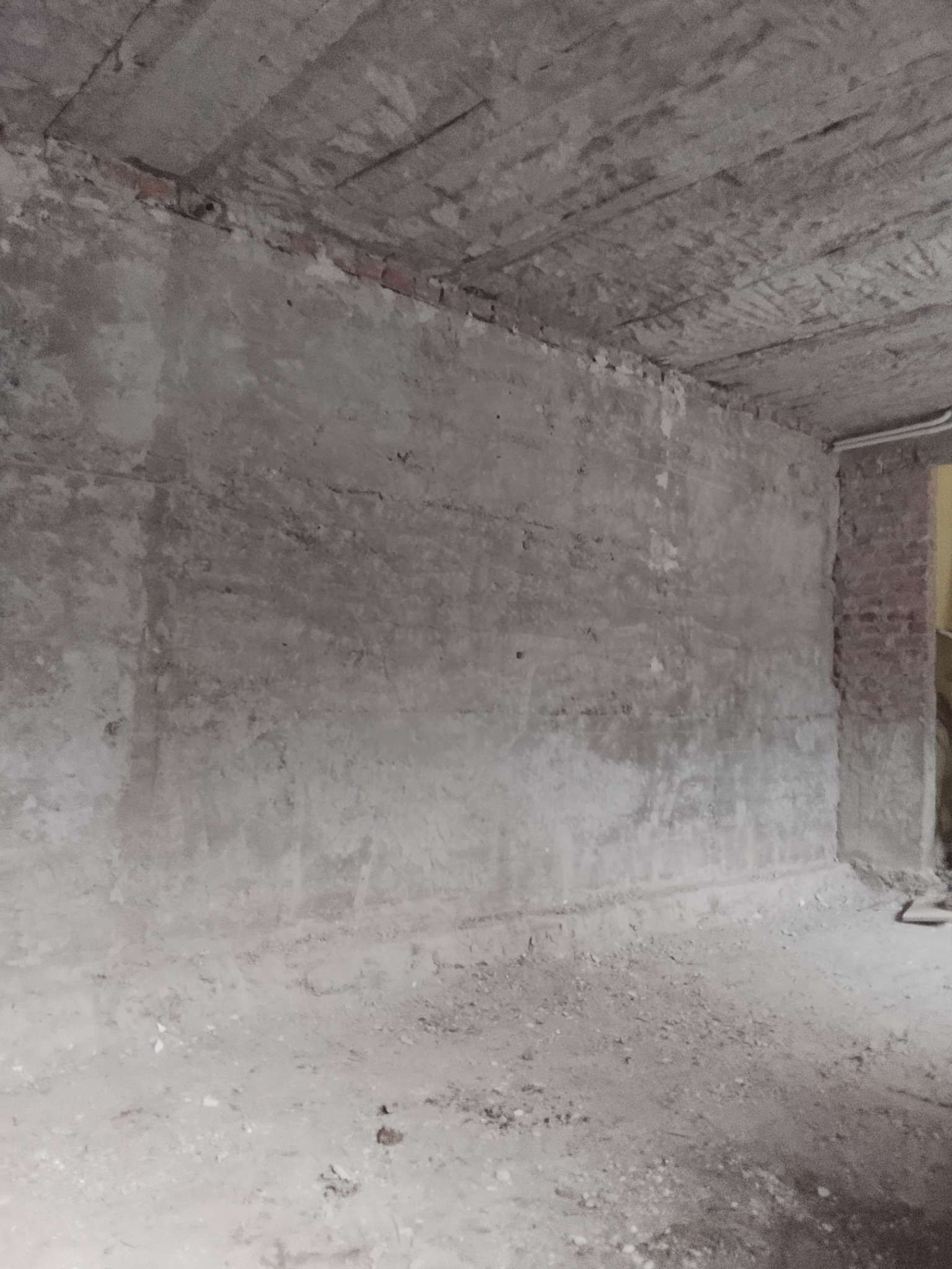 Surowe wnętrze z betonowym sufitem i ścianami w trakcie prac wykończeniowych. Widoczne ślady po instalacjach i niedoskonałości powierzchni. Podłoga pokryta gruzem.