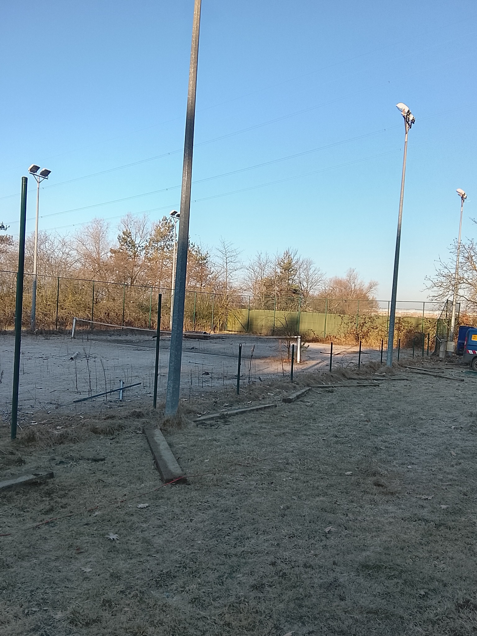 Zdemontowane ogrodzenie boiska sportowego w zimowej scenerii. Widoczne słupy oświetleniowe i fragment pojazdu w tle. Teren pokryty szronem.