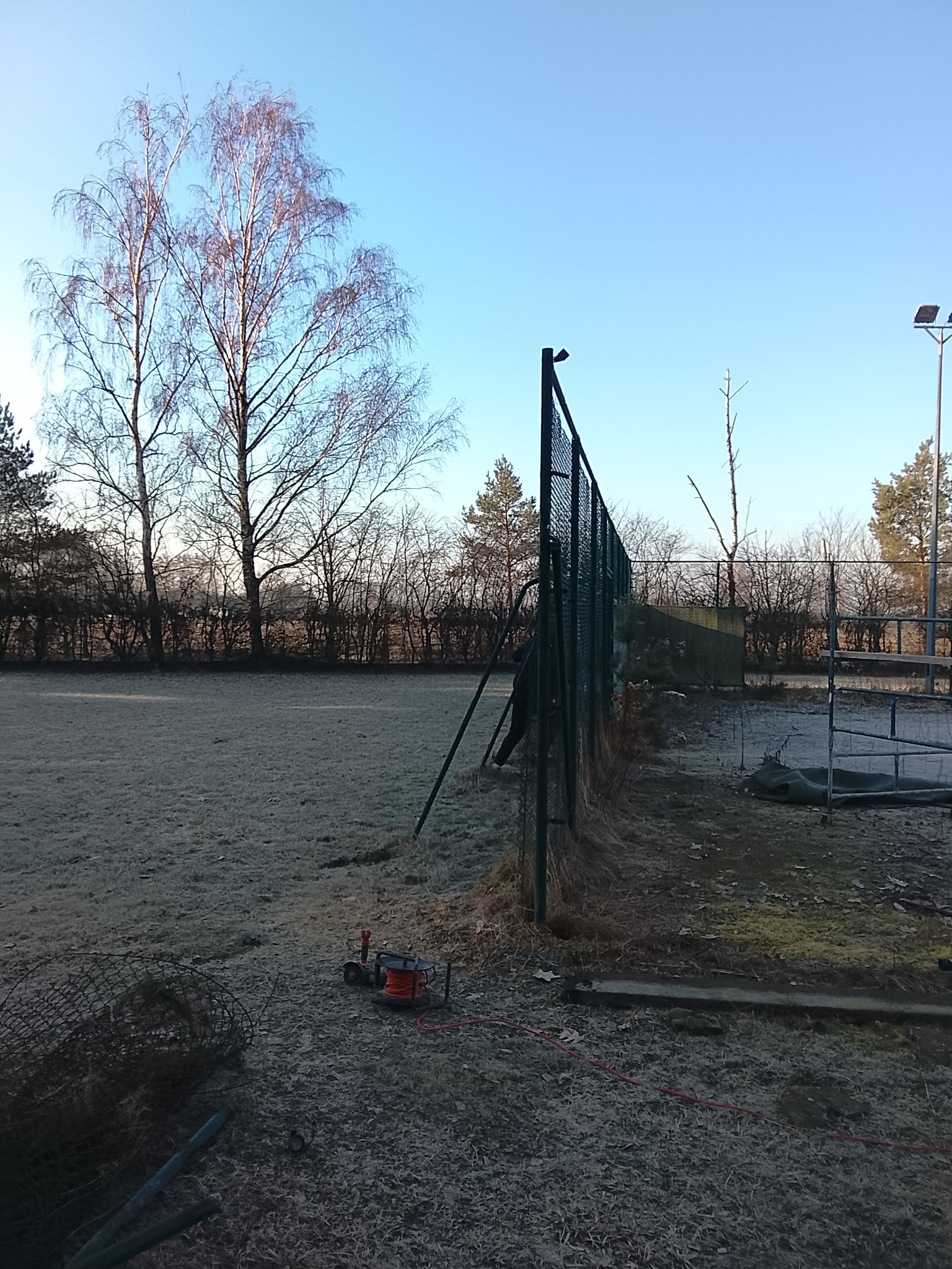 Montaż siatki ogrodzeniowej na zewnątrz w mroźny dzień. Widoczne narzędzia i materiały budowlane. Praca w toku, teren przygotowany pod ogrodzenie.