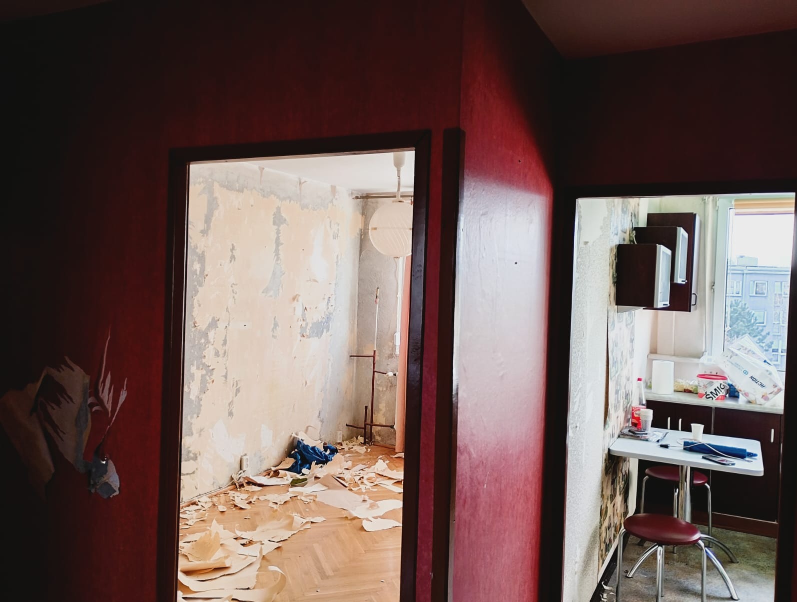 Pokój w trakcie remontu: zerwane tapety, widoczne warstwy farby, resztki materiałów na podłodze. Czerwone ściany w sąsiednim pomieszczeniu. Widok przez dwa otwarte przejścia.