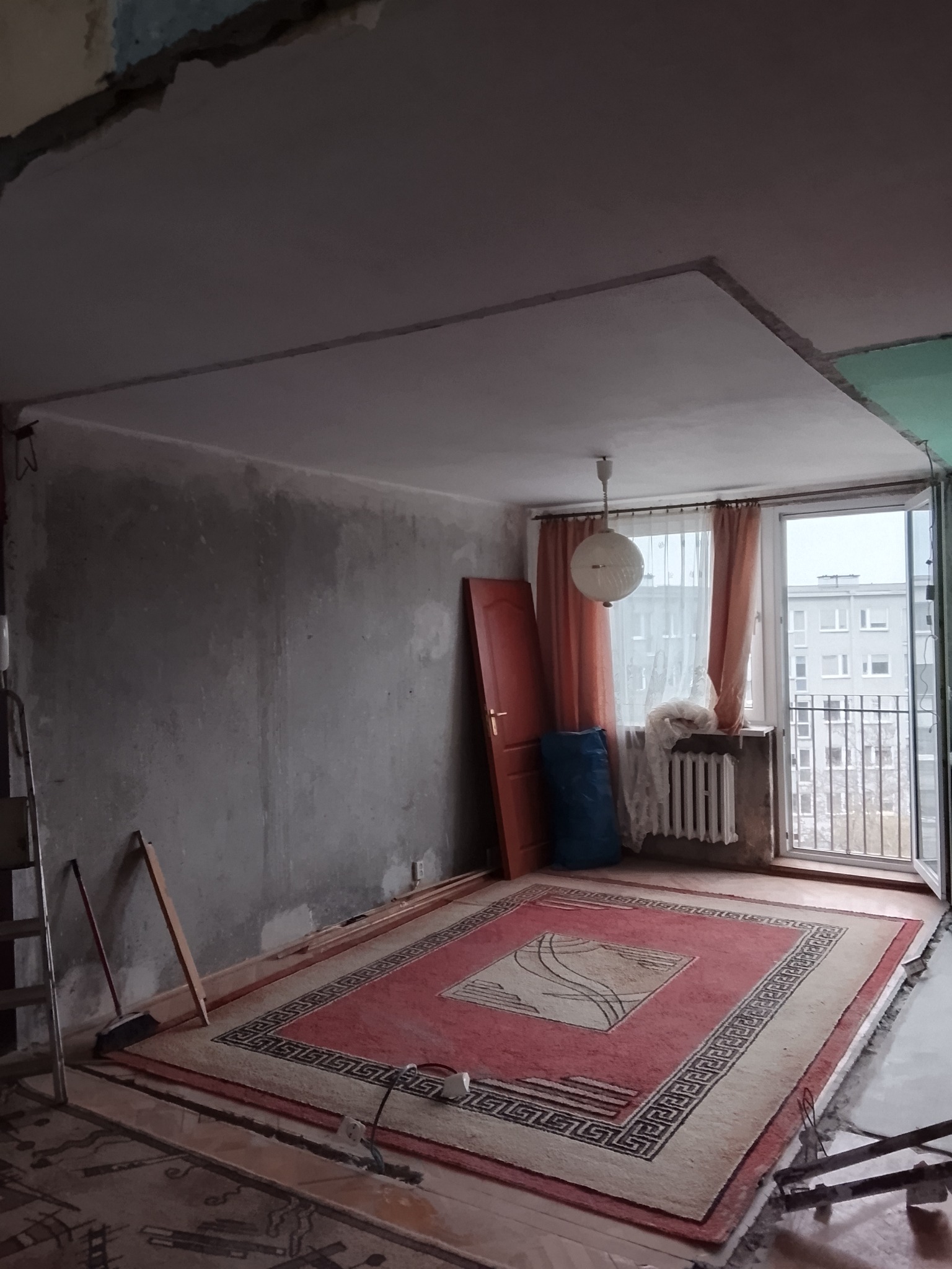 Pokój w trakcie remontu: surowe ściany, odsłonięty sufit, dywan zabezpieczony folią, drabina oparta o ścianę. Widok na balkon i budynek za oknem.