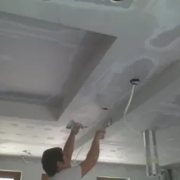 Pracownik wygładza szpachlą łączenia płyt gipsowo-kartonowych na suficie podwieszanym z wnękami, widoczne przewody elektryczne i metalowa rura wentylacyjna.