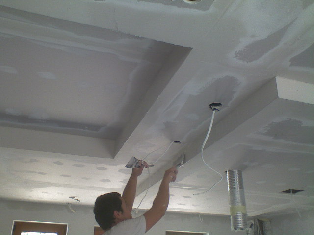 Pracownik wygładza szpachlą łączenia płyt gipsowo-kartonowych na suficie podwieszanym z wnękami, widoczne przewody elektryczne i metalowa rura wentylacyjna.