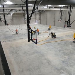 STALLION EPOXY - Prace na posadzce hali sportowej z koszami. Szlifowanie betonu z użyciem maszyn i odkurzaczy przemysłowych. Dwóch pracowników w odblaskowych ubraniach.