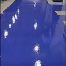 STALLION EPOXY - Błyszcząca, niebieska posadzka przemysłowa z żółtymi liniami oddzielającymi strefy w hali produkcyjnej. Gładka powierzchnia odbija światło, podkreślając czystość.