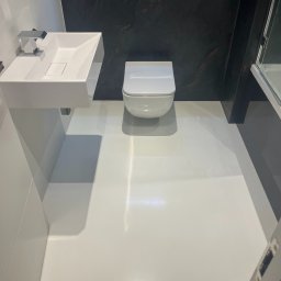 STALLION EPOXY - Biała, lśniąca posadzka w nowoczesnej łazience z wiszącą toaletą i umywalką. Ciemna ściana z efektem marmuru dodaje elegancji. Minimalistyczny design.