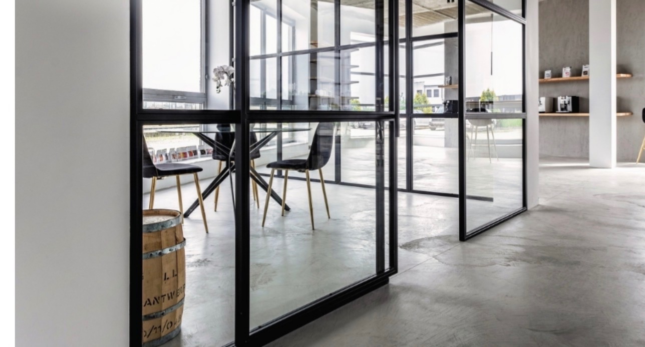 Nowoczesne biuro z posadzką mikrocementową, szklane ściany z czarnymi ramami, drewniana beczka w rogu. Minimalistyczny design, szara kolorystyka.