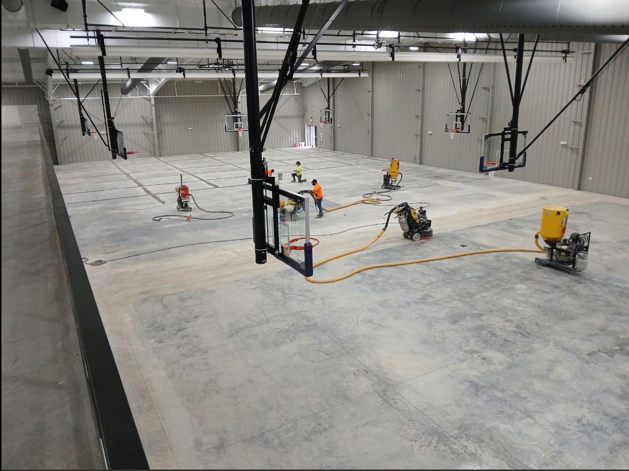 Prace na posadzce hali sportowej z koszami. Szlifowanie betonu z użyciem maszyn i odkurzaczy przemysłowych. Dwóch pracowników w odblaskowych ubraniach.