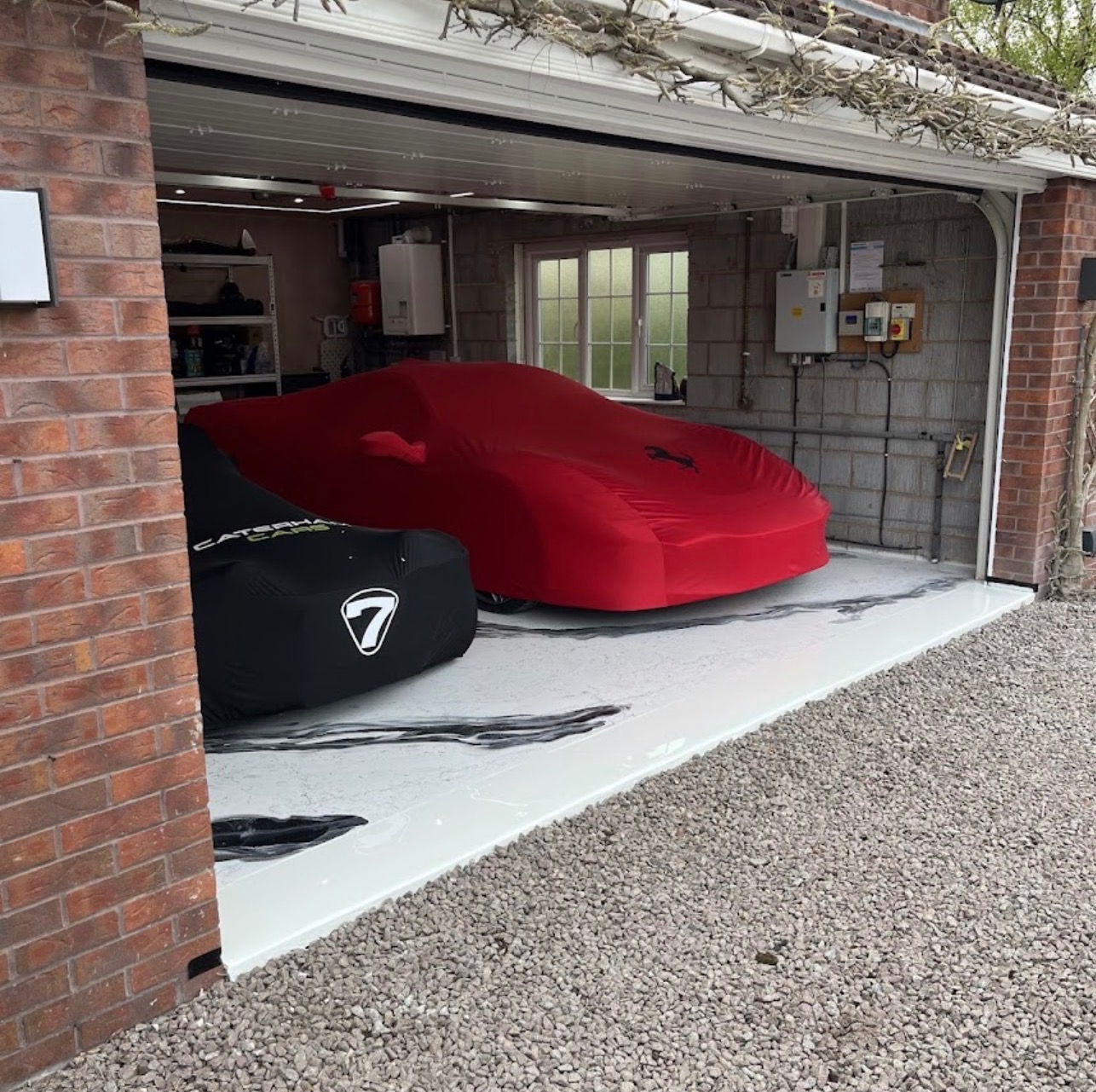 Biała posadzka w garażu z dwoma samochodami pod pokrowcami, jeden czerwony z logo Ferrari, drugi czarny z logo Caterham Cars, szara ściana i okno w tle.