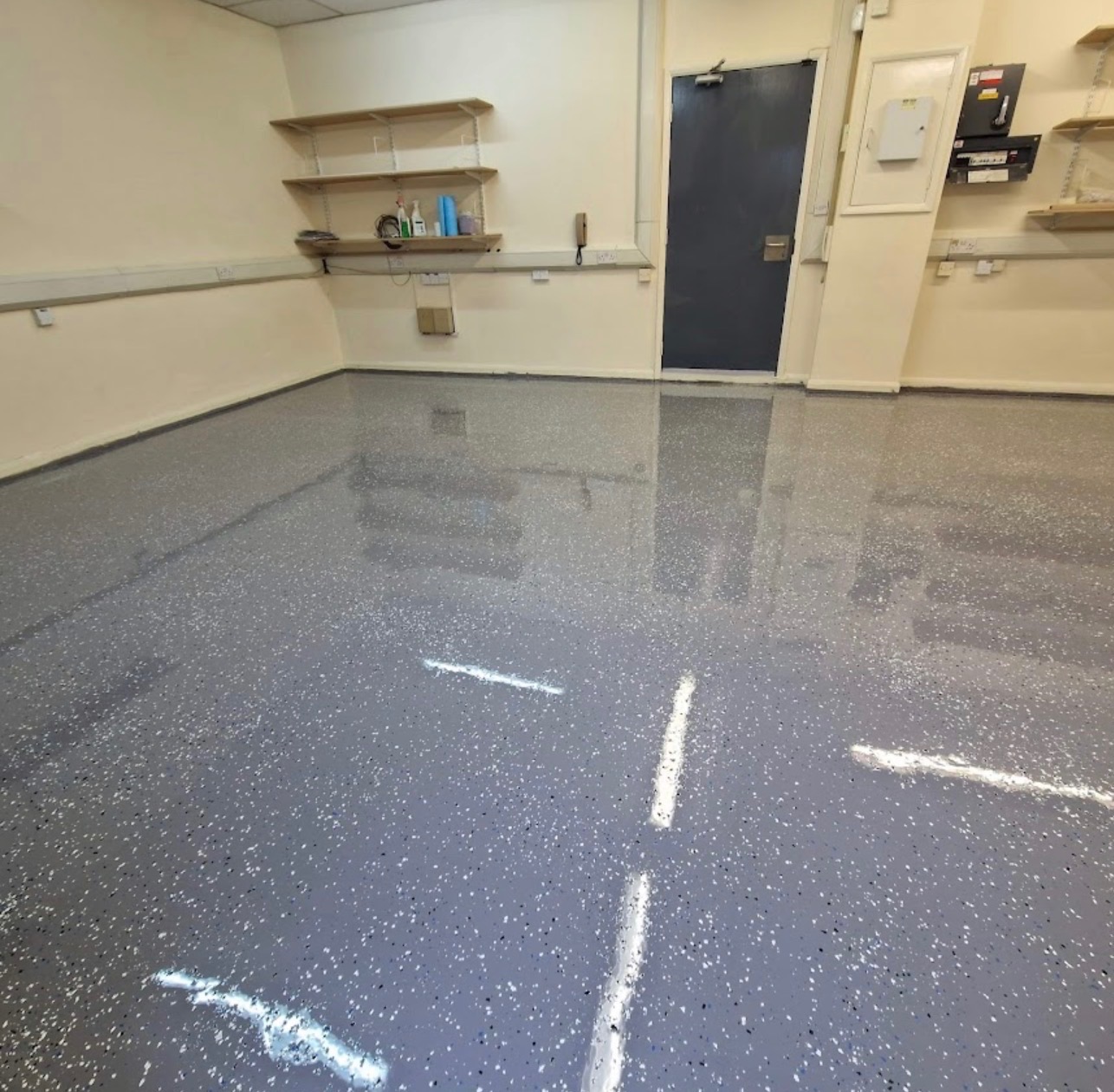 Szara, błyszcząca posadzka z drobnymi, kolorowymi wtrąceniami w jasnym pomieszczeniu z półkami i szarą ścianą. Widoczne odbicia światła na powierzchni podłogi.