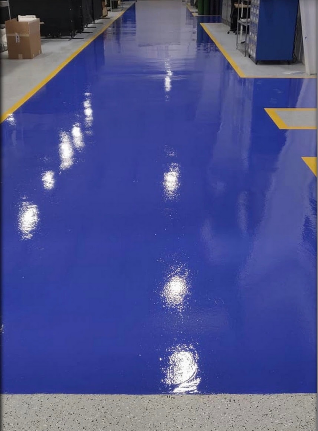 Błyszcząca, niebieska posadzka przemysłowa z żółtymi liniami oddzielającymi strefy w hali produkcyjnej. Gładka powierzchnia odbija światło, podkreślając czystość.