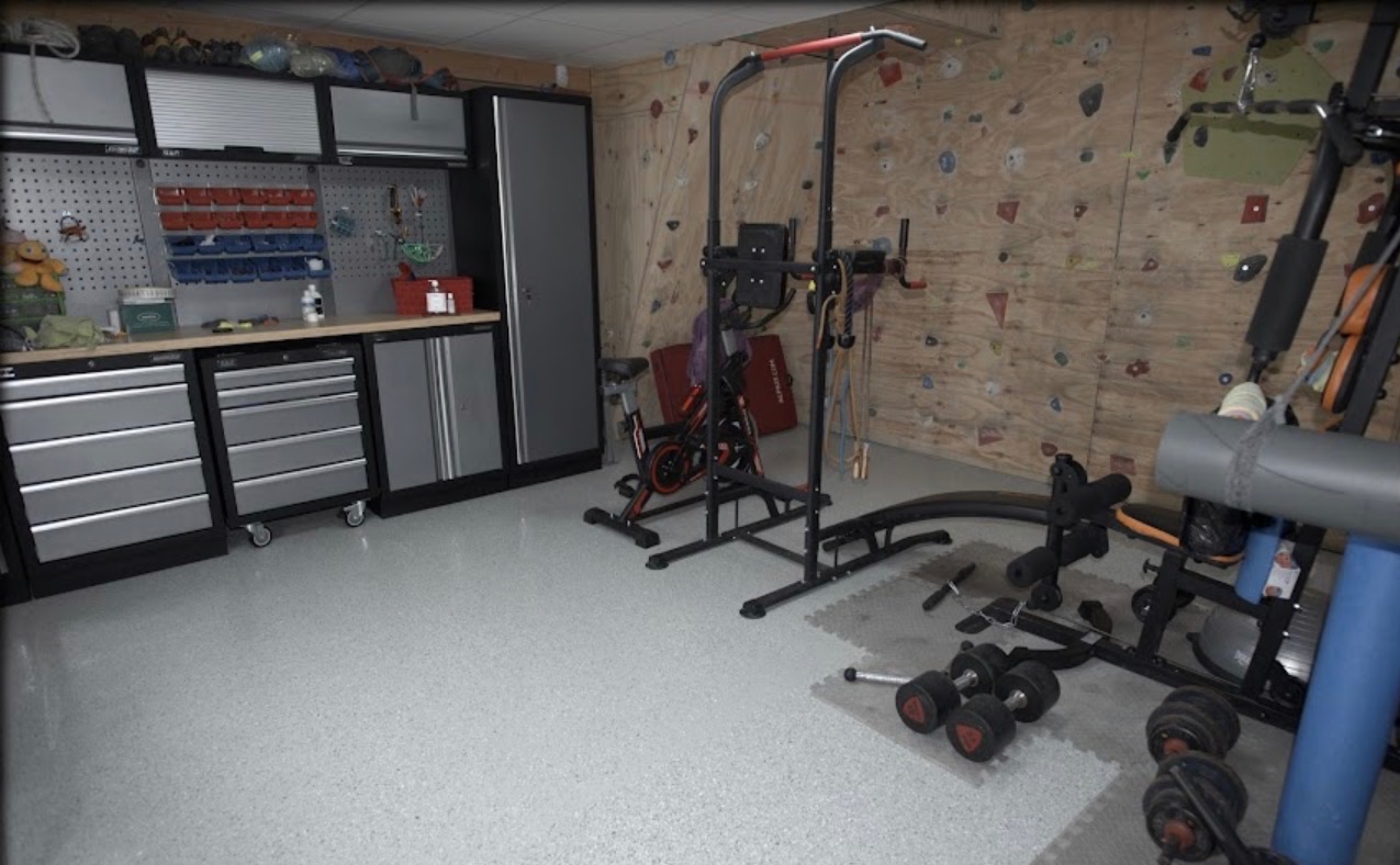 Jasnoszara, nakrapiana podłoga w domowej siłowni z wyposażeniem: szafki narzędziowe, drabinka do wspinaczki, rower stacjonarny i ciężary. Nowoczesny design.