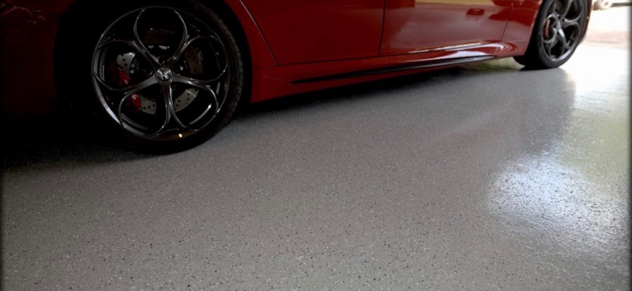 Szara, błyszcząca posadzka z drobnymi plamkami, obok czerwony samochód Alfa Romeo z widocznym kołem. Gładka powierzchnia, garaż.