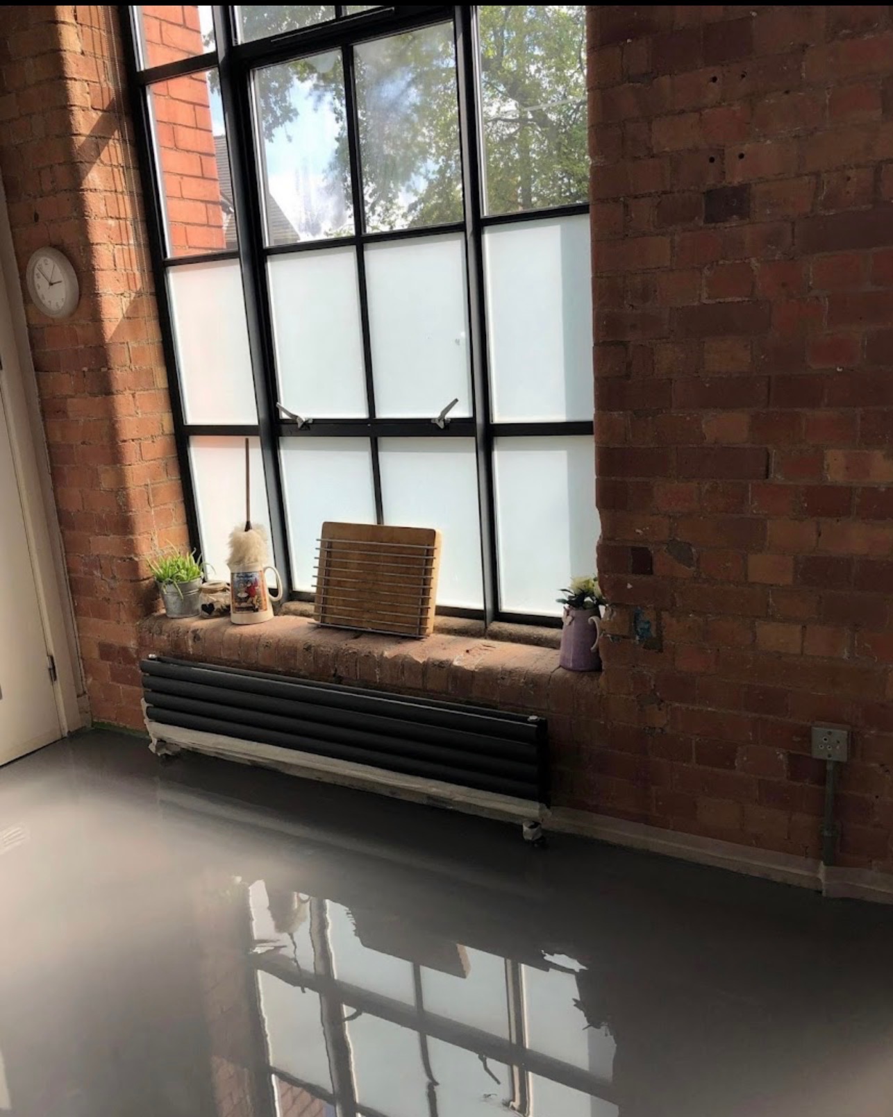 Szara, błyszcząca posadzka żywiczna w loftowym wnętrzu z ceglaną ścianą i dużym oknem. Widoczne odbicie okna w posadzce. Grzejnik pod oknem.