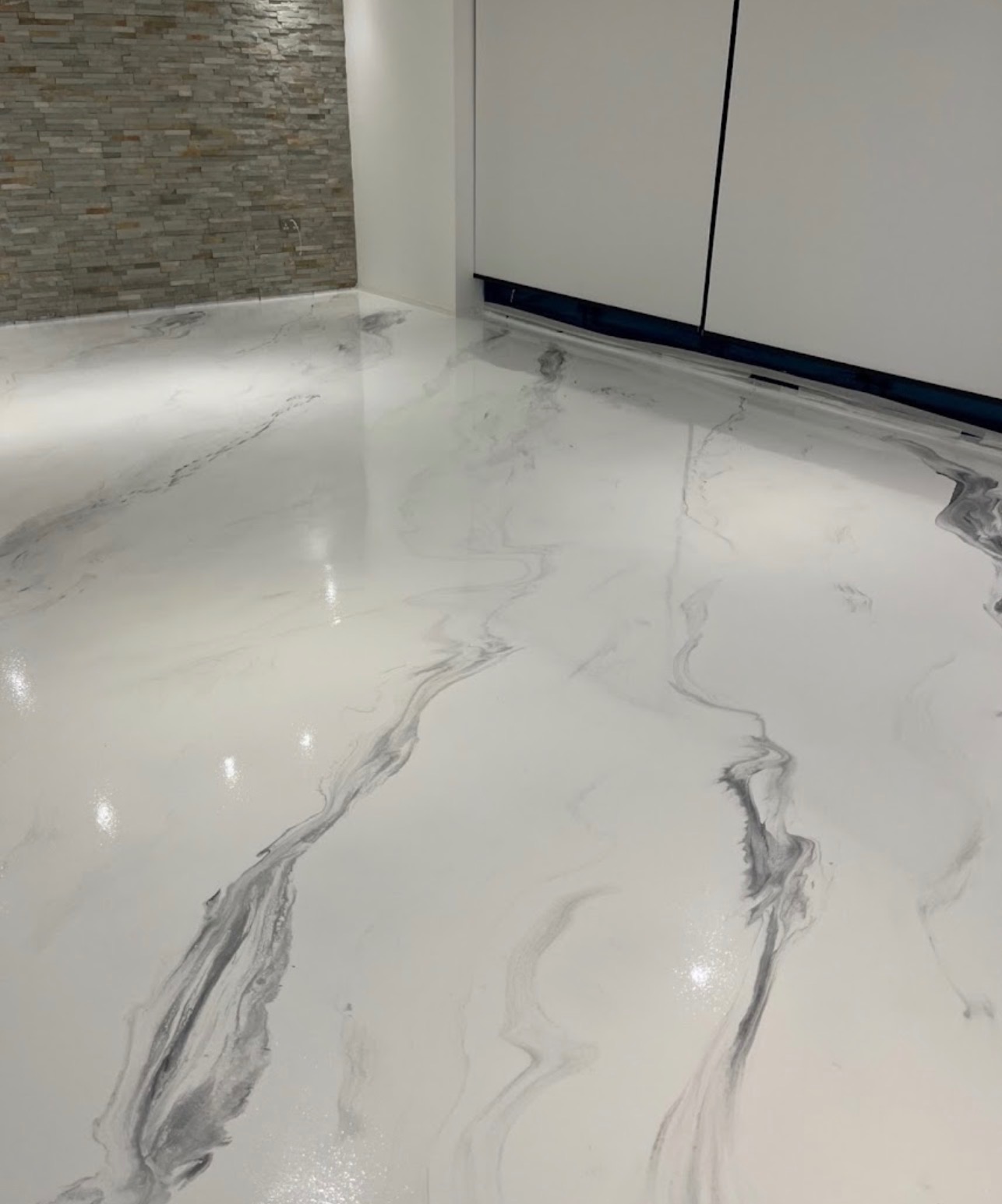 Błyszcząca, biała posadzka z szarymi żyłkami imitującymi marmur w nowoczesnym wnętrzu z kamienną ścianą i białymi szafkami. Gładka powierzchnia odbija światło.
