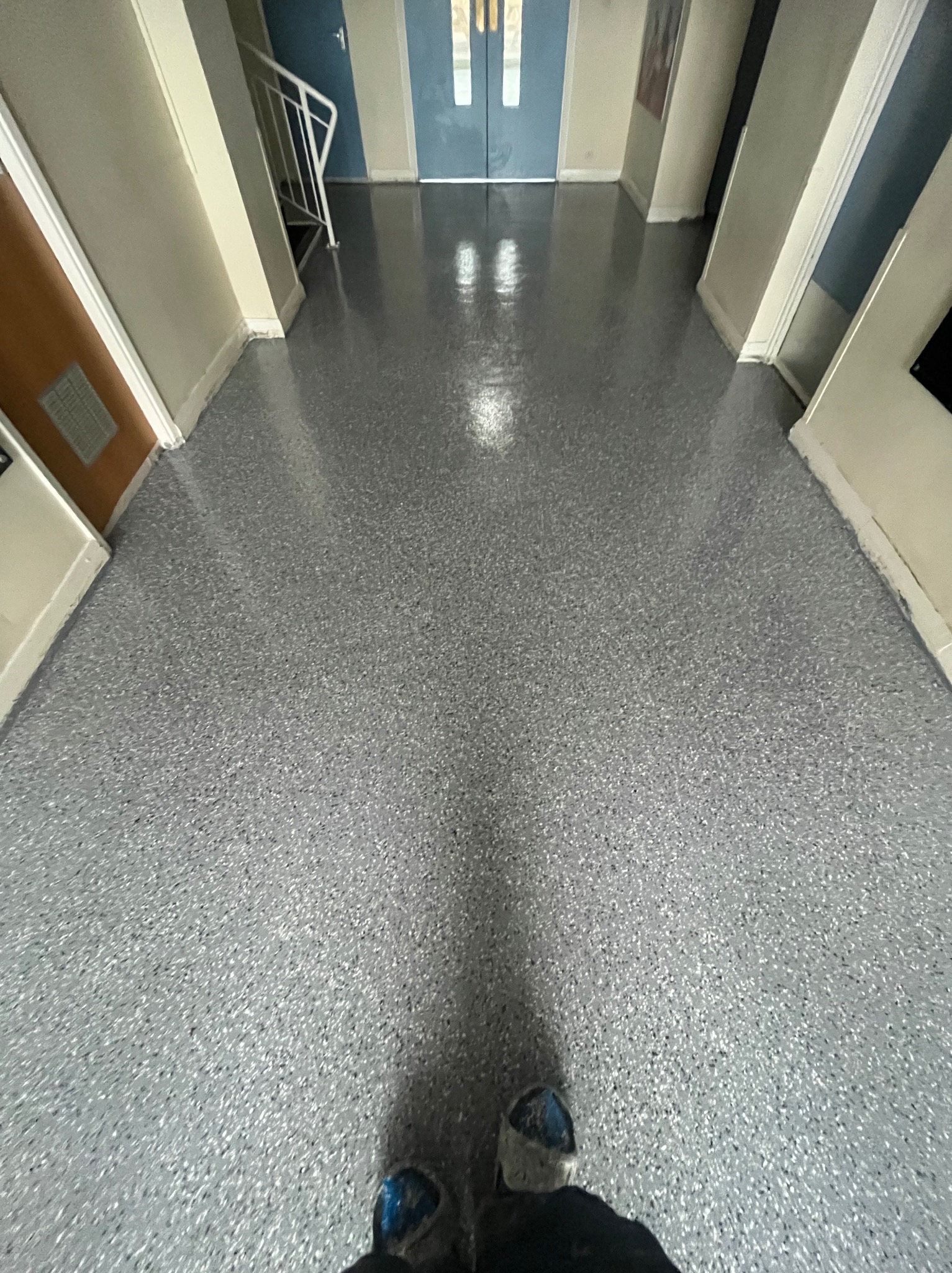 Błyszcząca, szara posadzka z drobnymi, ciemnymi inkluzjami w korytarzu. Widoczne drzwi i fragment schodów w tle. Perspektywa z dołu, z butami w kadrze.