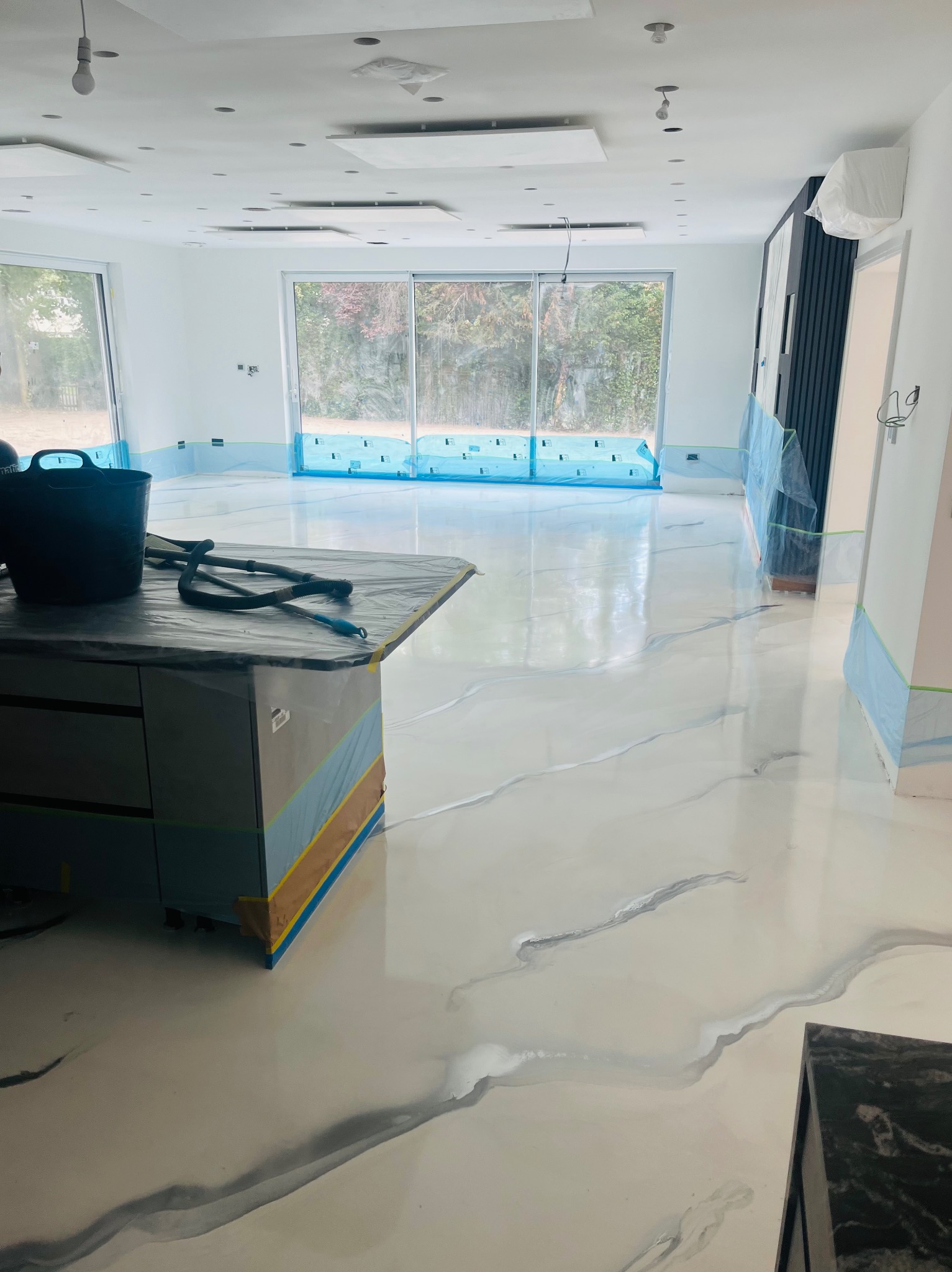 Jasna, błyszcząca posadzka z szarymi wzorami imitującymi marmur w przestronnym, jasnym wnętrzu. Widoczne zabezpieczenia folią i taśmą malarską.