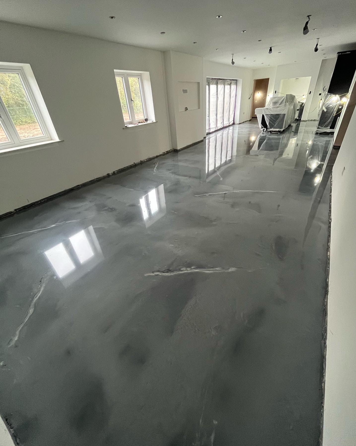 Błyszcząca, szara posadzka z efektem marmuru w jasnym wnętrzu. Widoczne okna i częściowo zakryte meble. Nowoczesny design podłogi z refleksami światła.