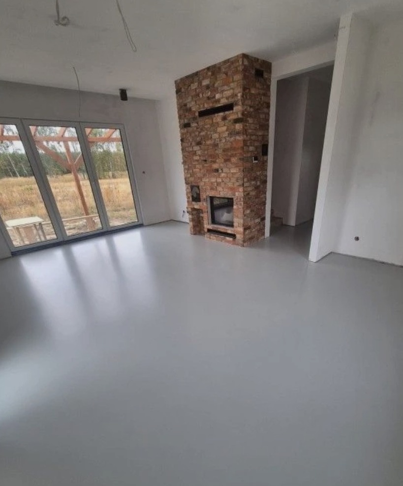 Jasnoszara, gładka posadzka w surowym wnętrzu z ceglanym kominkiem i dużym oknem tarasowym. Minimalistyczny design, widoczne niedokończone wykończenia ścian.
