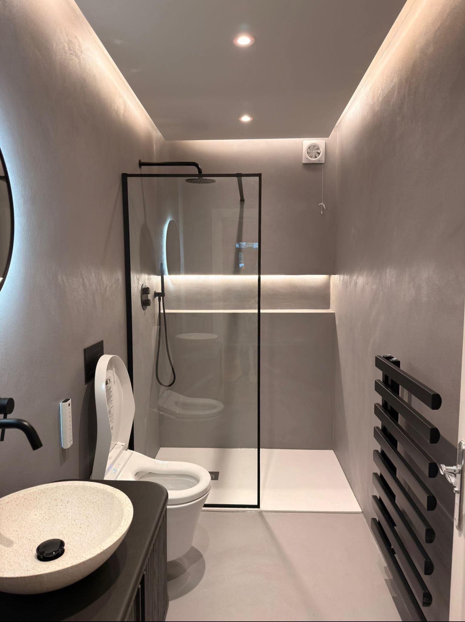 Elegancka łazienka z kabiną prysznicową typu walk-in, umywalką nablatową i grzejnikiem drabinkowym. Ściany wykończone w szarym odcieniu z efektem dekoracyjnym.