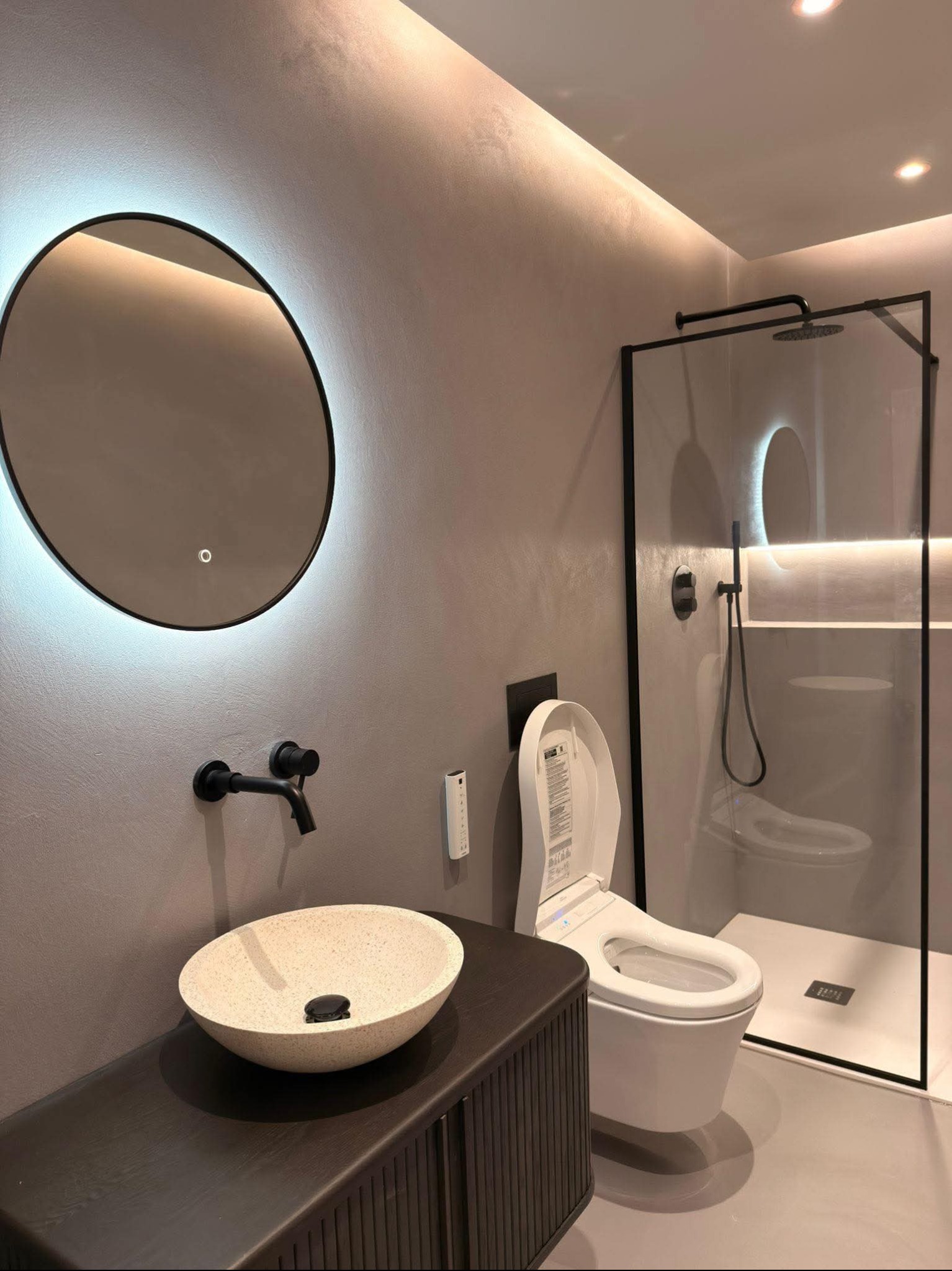 Stylowa łazienka z szarą ścianą wykończoną mikrocementem, okrągłym lustrem z podświetleniem LED, kamienną umywalką i kabiną prysznicową z czarnymi ramami.