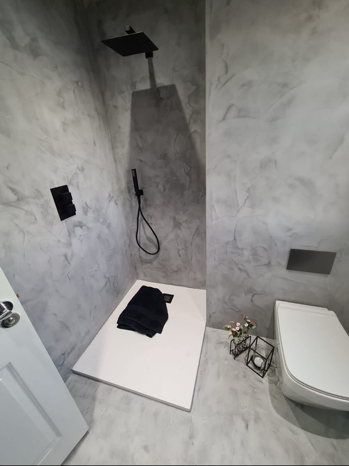 Nowoczesna łazienka z kabiną prysznicową typu walk-in, wykończona szarą, wzorzystą powłoką na ścianach i podłodze, imitującą kamień. Czarna armatura i biała ceramika.