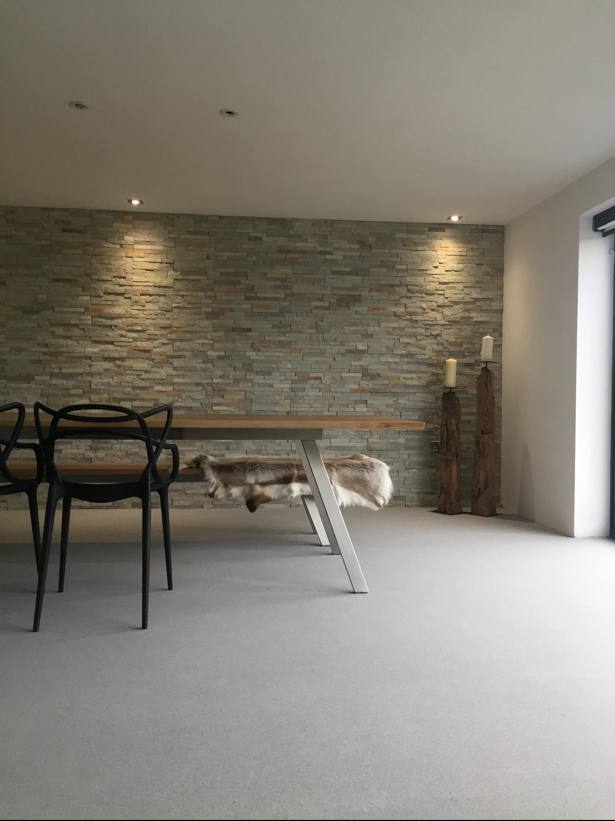 Szara, gładka posadzka w nowoczesnym wnętrzu z kamienną ścianą i drewnianym stołem. Minimalistyczny design z akcentami naturalnych materiałów.