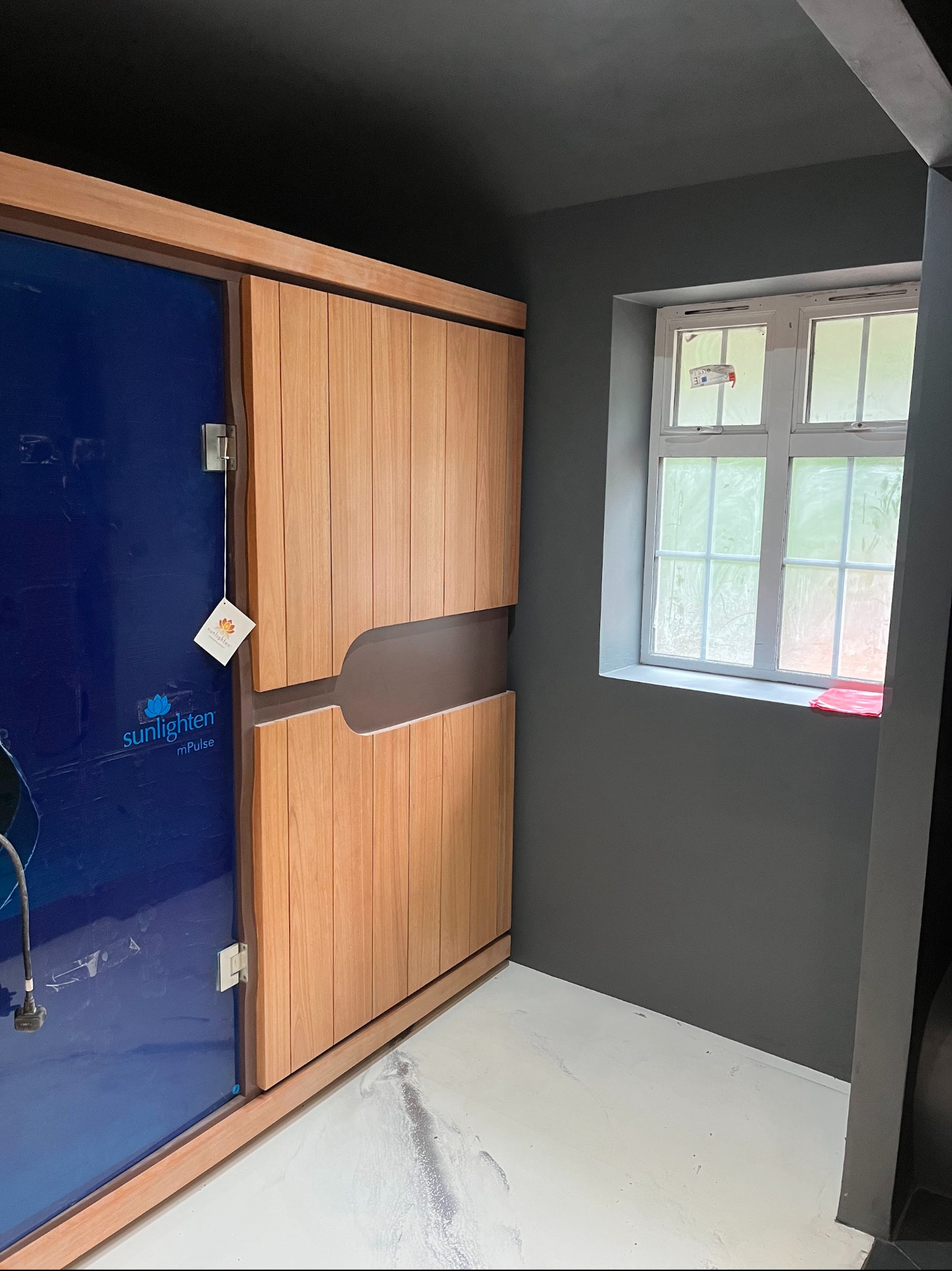 Nowoczesna kabina infrared z drewnianymi drzwiami przesuwnymi i niebieską szybą, na tle szarej ściany i białej podłogi z delikatnym połyskiem, oświetlona naturalnym światłem z okna.