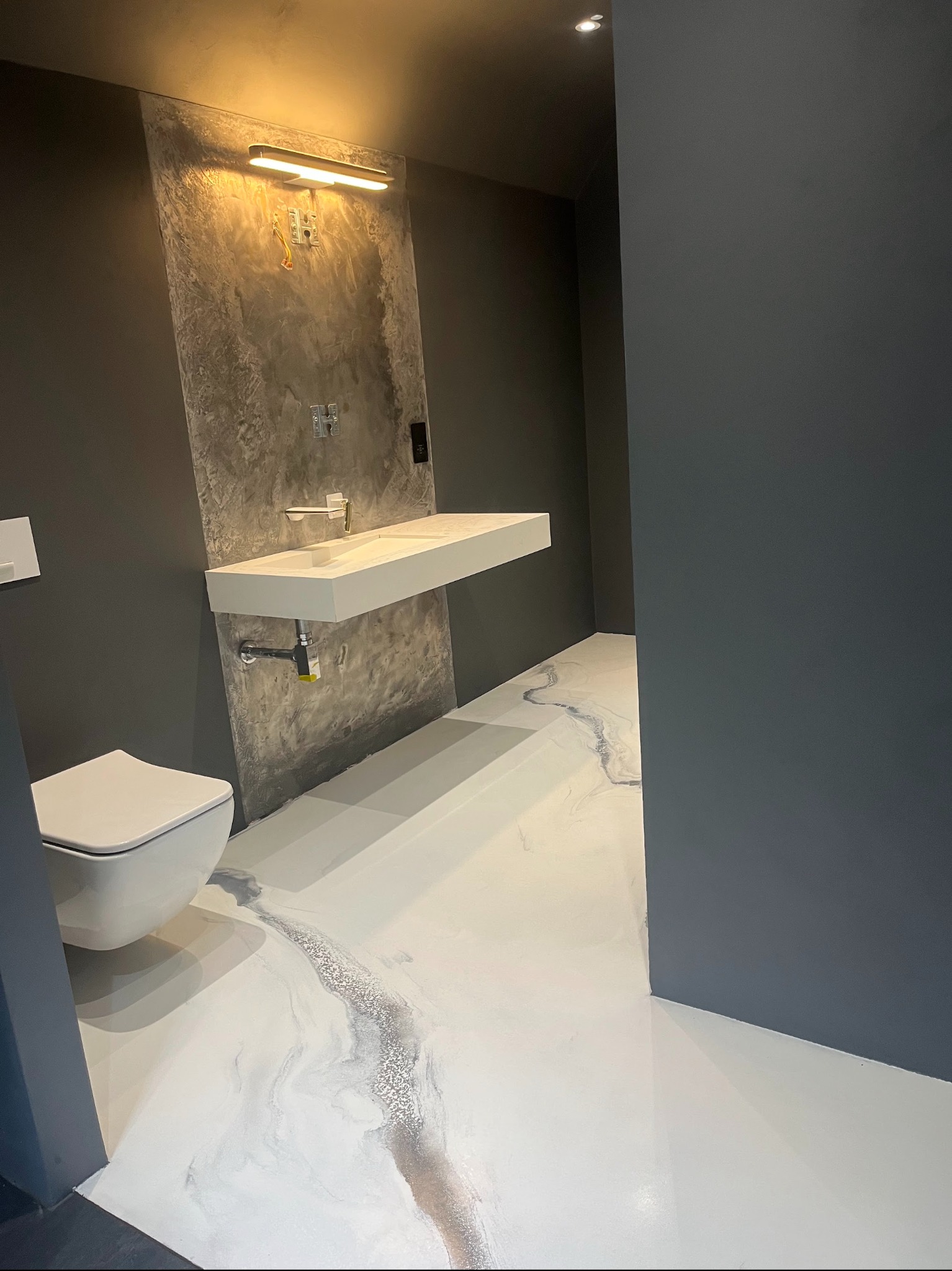 Biała posadzka z efektem marmuru w nowoczesnej łazience z szarą ścianą i podwieszaną umywalką. Widoczne detale wykończenia i oświetlenie.