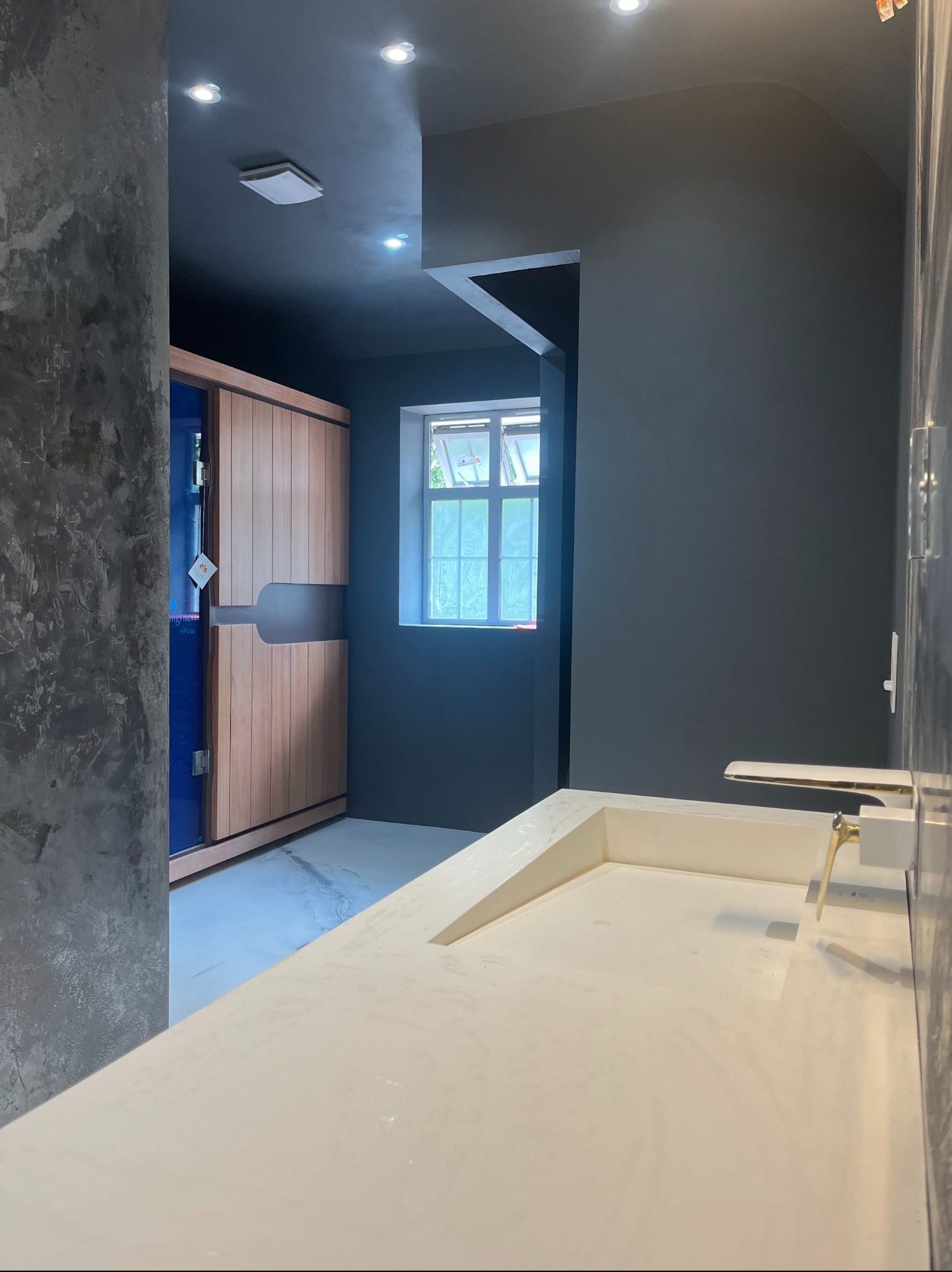 Nowoczesna łazienka z posadzką żywiczną, drewnianą sauną i umywalką z blatem z mikrocementu. Szare ściany i sufit, okno z widokiem na zieleń.