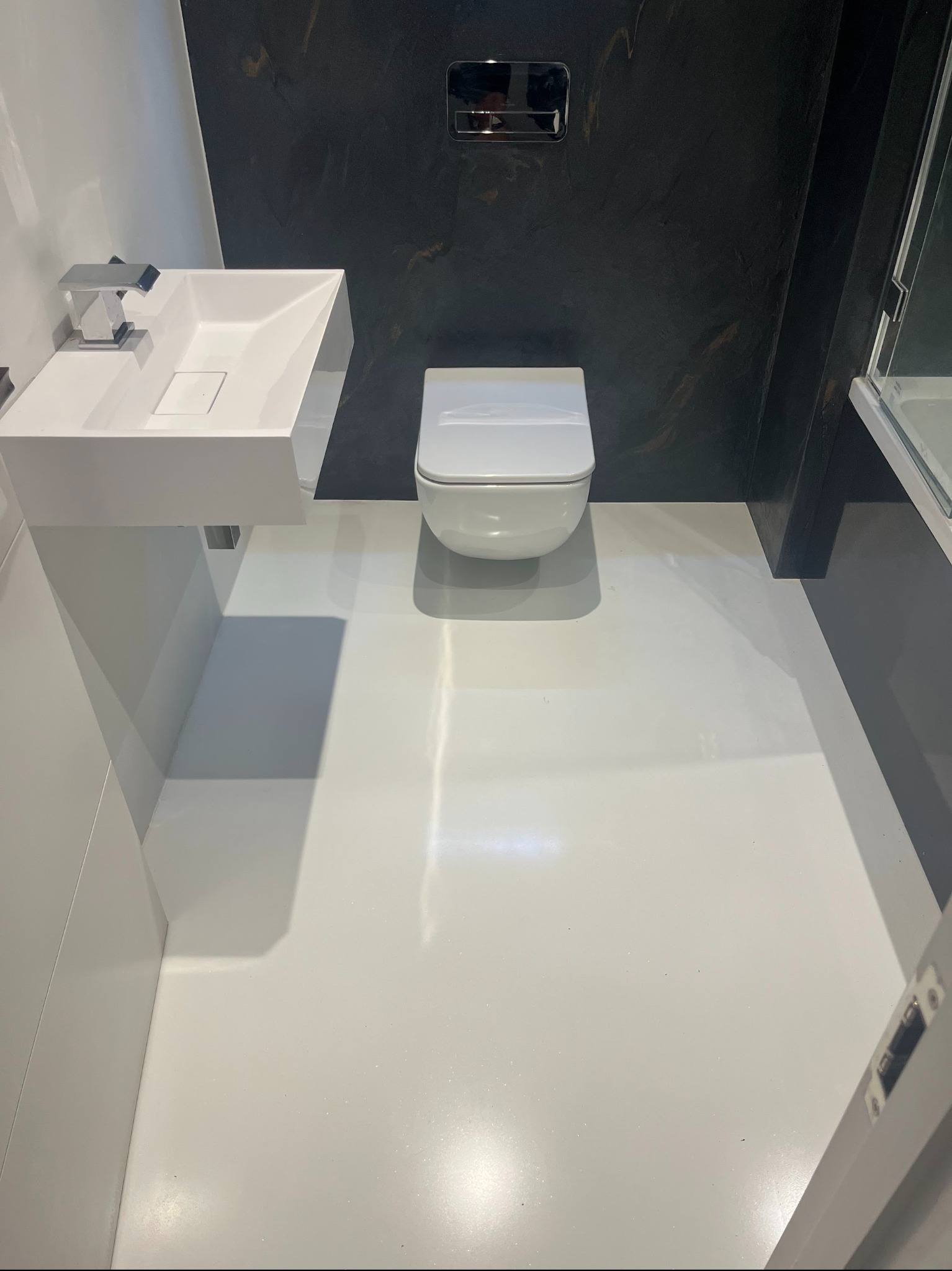 Biała, lśniąca posadzka w nowoczesnej łazience z wiszącą toaletą i umywalką. Ciemna ściana z efektem marmuru dodaje elegancji. Minimalistyczny design.